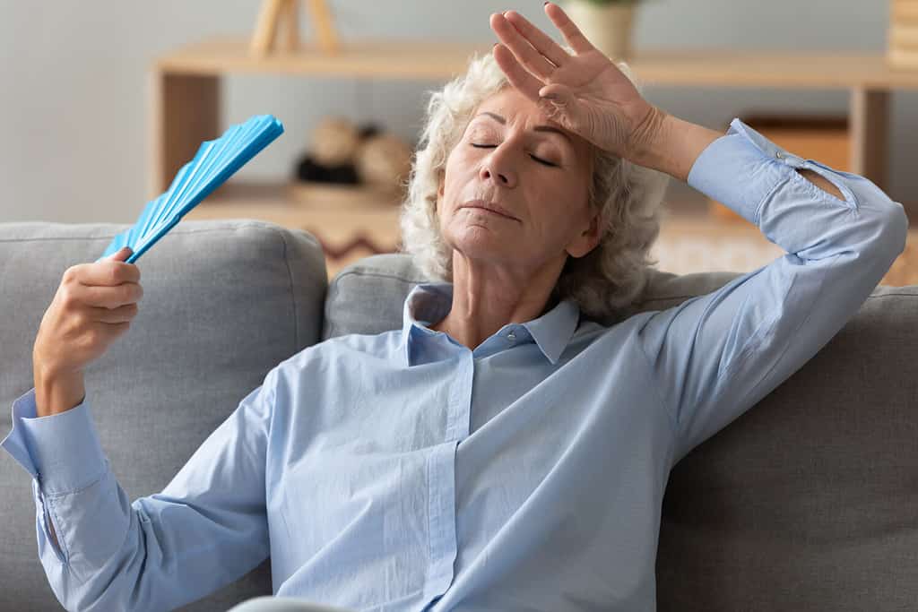 Tired annoyed senior woman holding waving fan feel hot sit on sofa at home without air conditioner, overheated exhausted old elder grandma sweating suffer from hormone problem summer heat concept