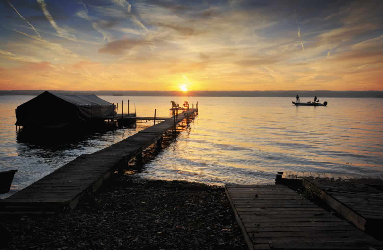 A beautiful autumn sunrise on the shores of Lake Cayuga in the Finger lakes region of New York state. A row boat is docked on the side of a pier. Two fisherman enjoy the sunrise from their boat.