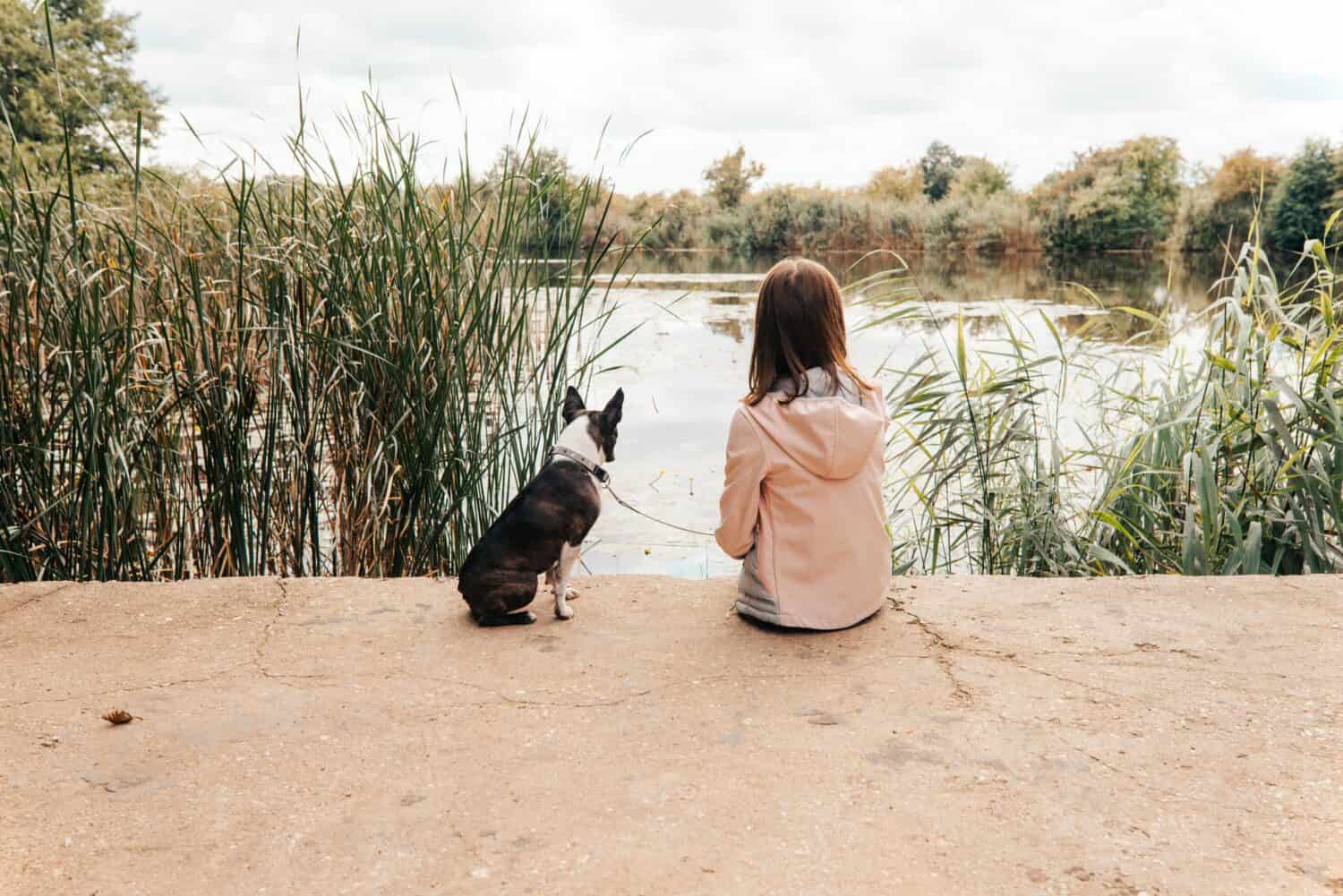 A little girl with a Boston Terrier sitting at a creek. 