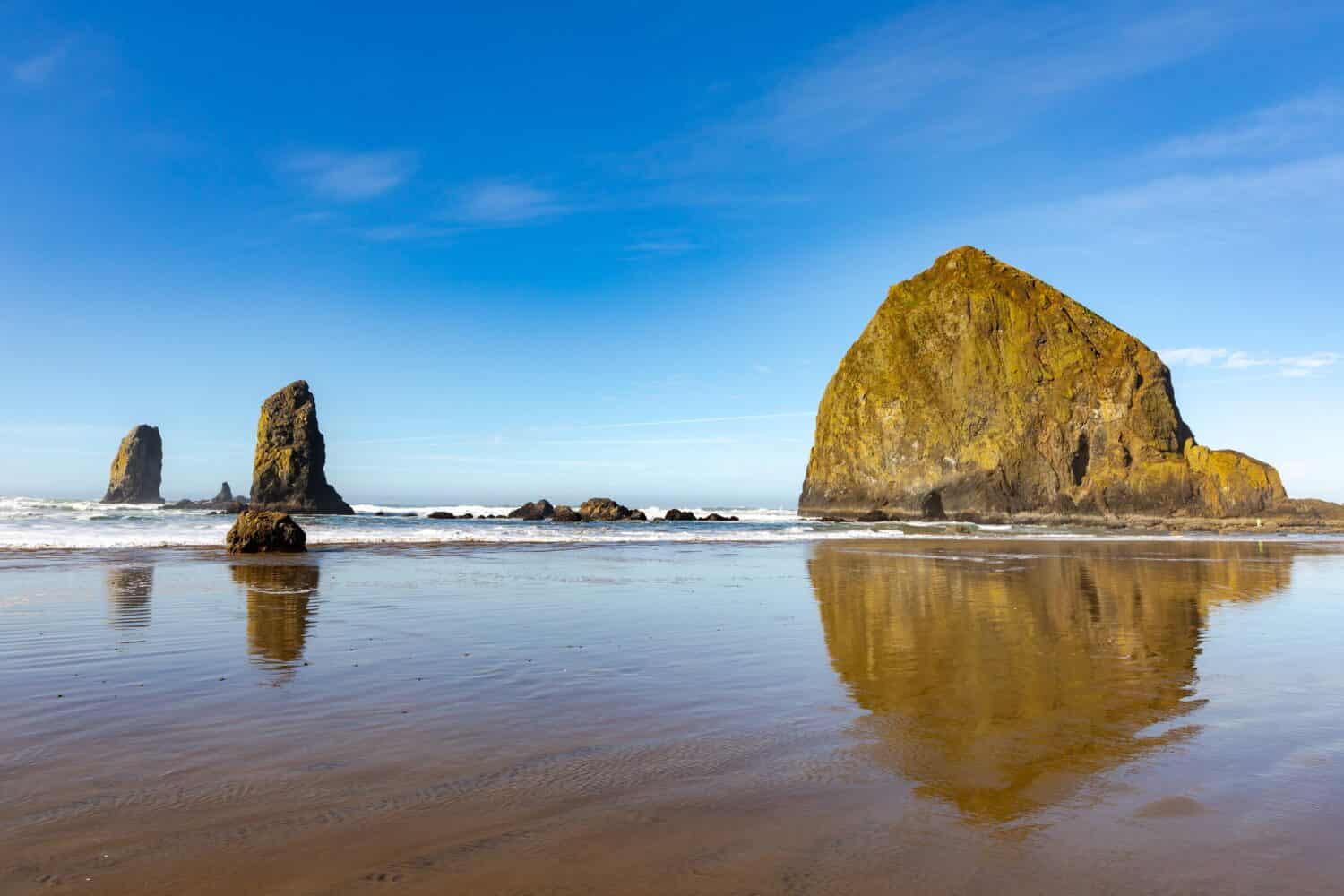 Haystack rock wide angle view with reflective water at coastline edge on blue sky sunny afternoon.