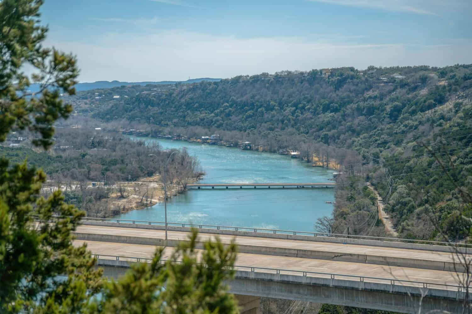 View from Lake Austin Dam of the Colorado River with concrete road bridge. Aerial landscape of water and roadway amid lush foliage of trees and sky on a sunny day.