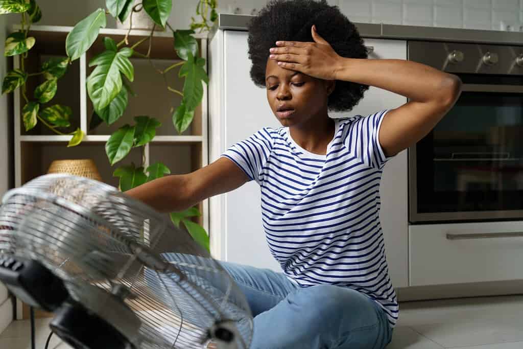 Unhappy African woman touching forehead having hot flashes using electric fan to cool off, overheated black female sitting in front of air cooler at home, surviving heatwave without air conditioning