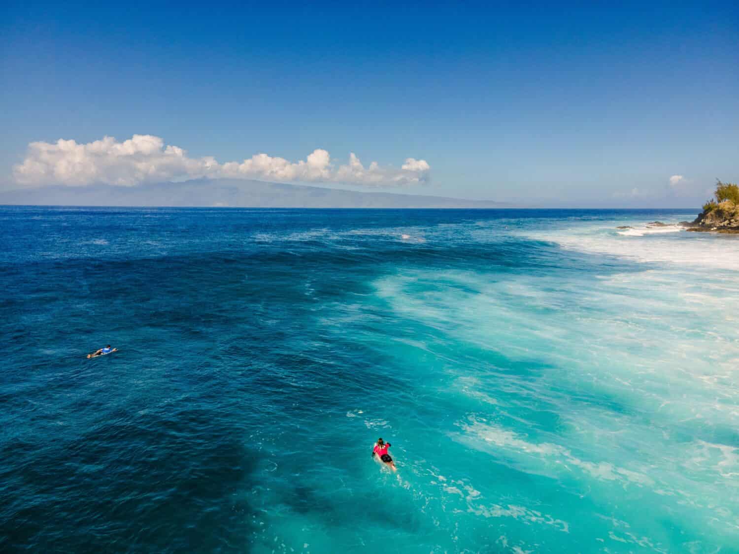 A surfer lying on a surfboard in the clear blue waters of Honolua Bay, Maui, HI.