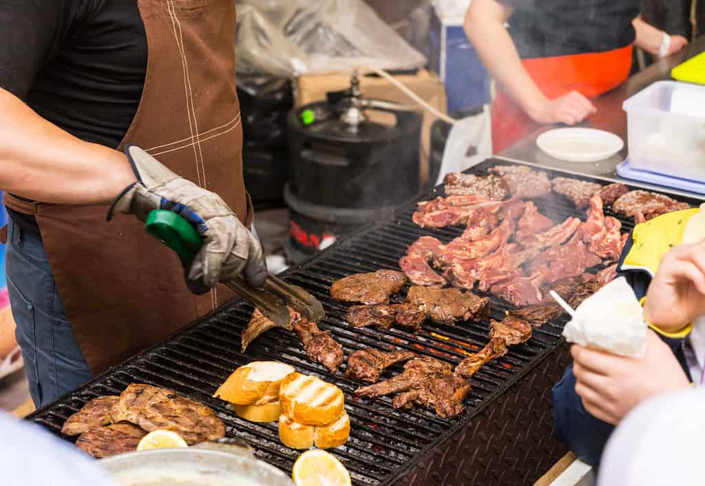 Person barbecuing meat at a catered event with a close up view of the chefs hands and meat cooking on the grill over a fire