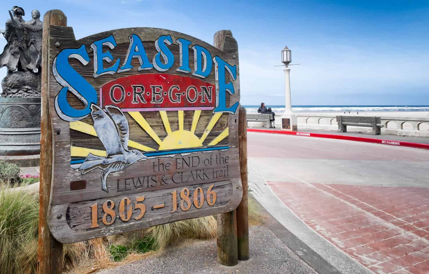 Seaside, Oregon sign commemorating its historic connection to the famous explorers Lewis & Clark, who ended their journey at this spot. A bronze statue of them and the ocean are in the background.