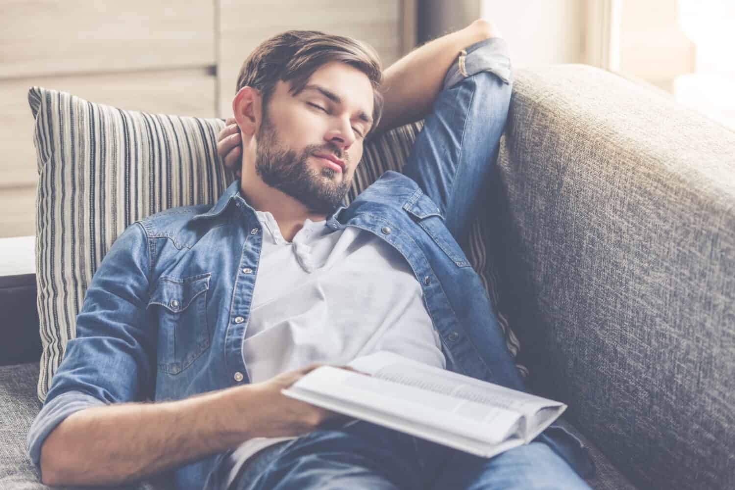 Handsome man is holding a book and napping while lying on couch at home