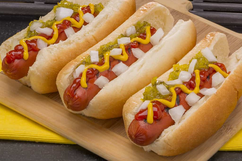 Three Hot Dogs with Mustard, Ketchup, pickle relish and onions on a wood cutting board