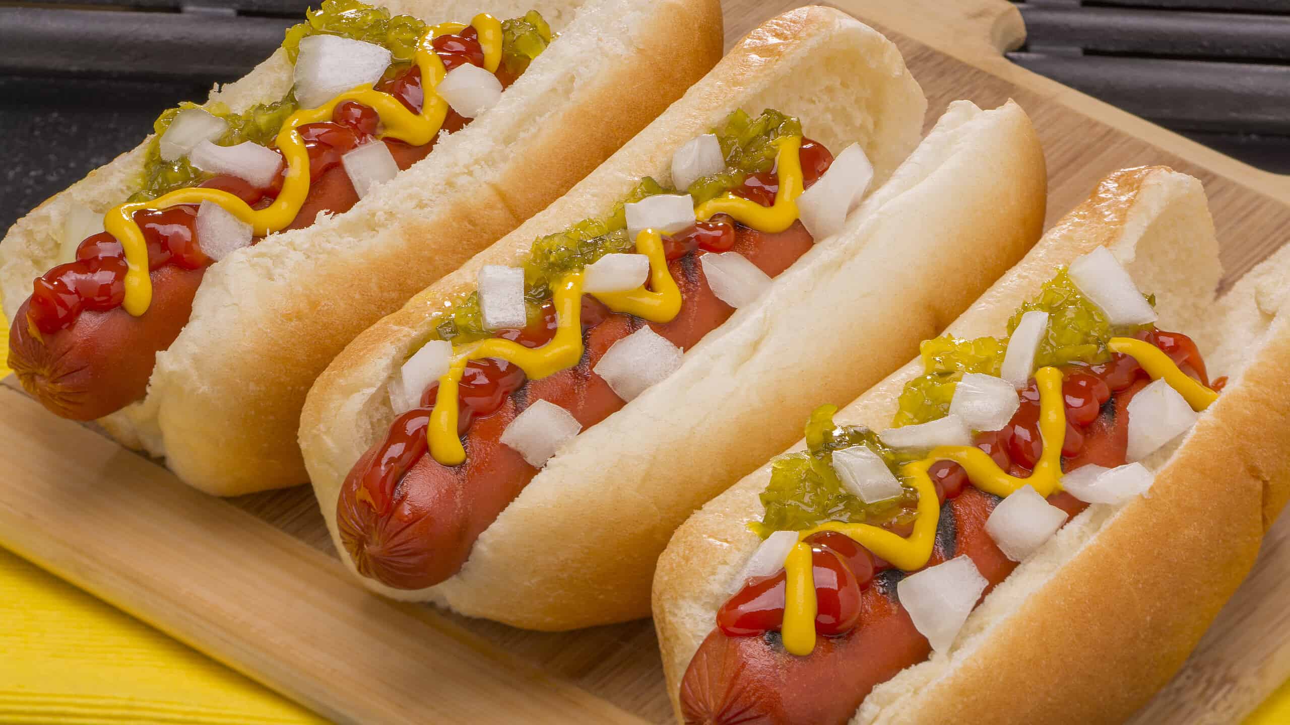 Three Hot Dogs with Mustard, Ketchup, pickle relish and onions on a wood cutting board