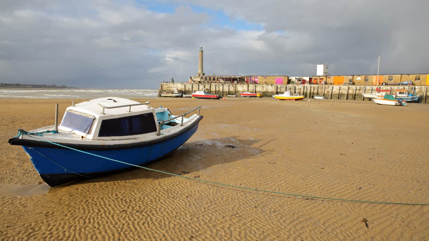 The beach at low tide with mooring boats and Margate Harbor Arm in the background, Margate, Kent, UK