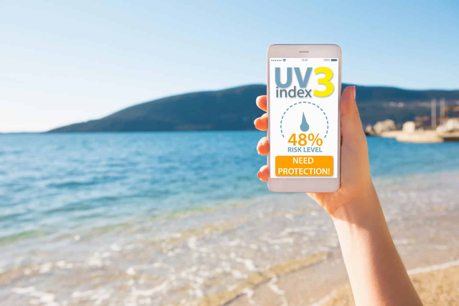 A woman is holding a phone. On the screen is a mobile application for determining the level of ultraviolet radiation. In the background the sea beach