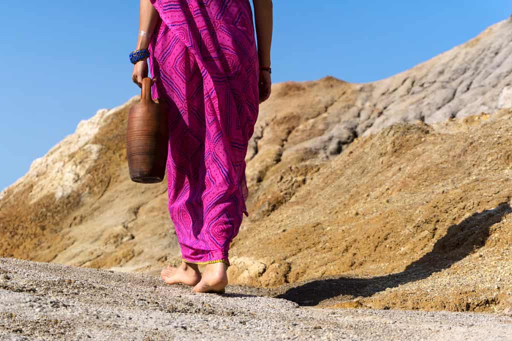 a barefooted woman in ethnical clothes carries in her hand a clay jug