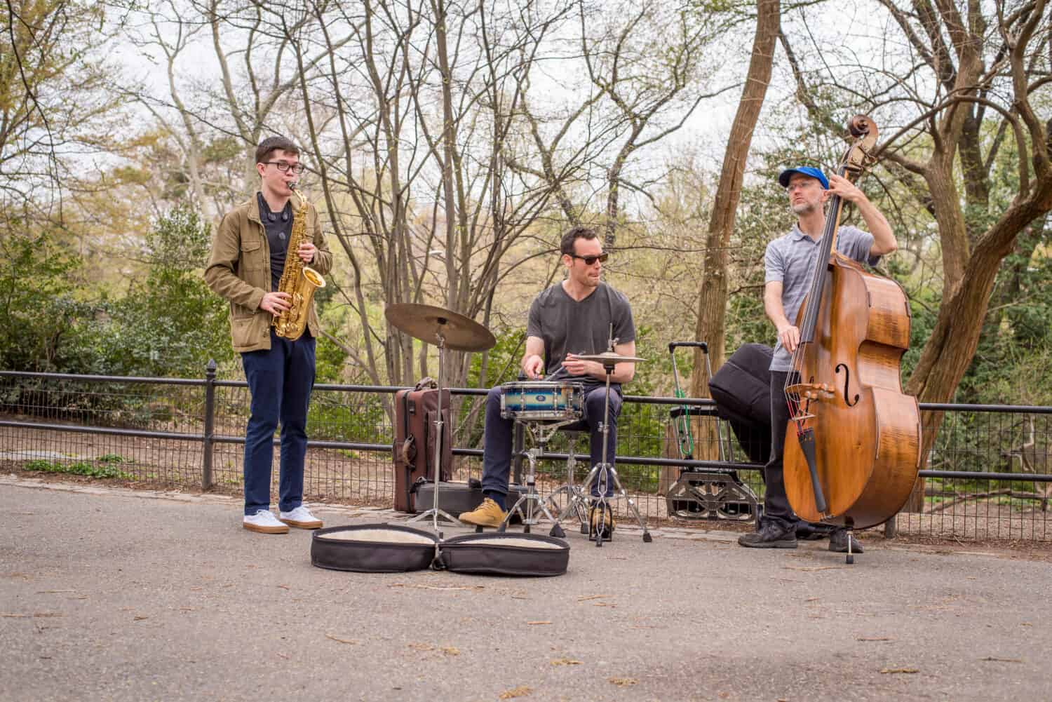 Jazz trio, alto saxophone, drums, and standup bass, play and busk in New York City's Central Park amongst the trees on the asphalt path