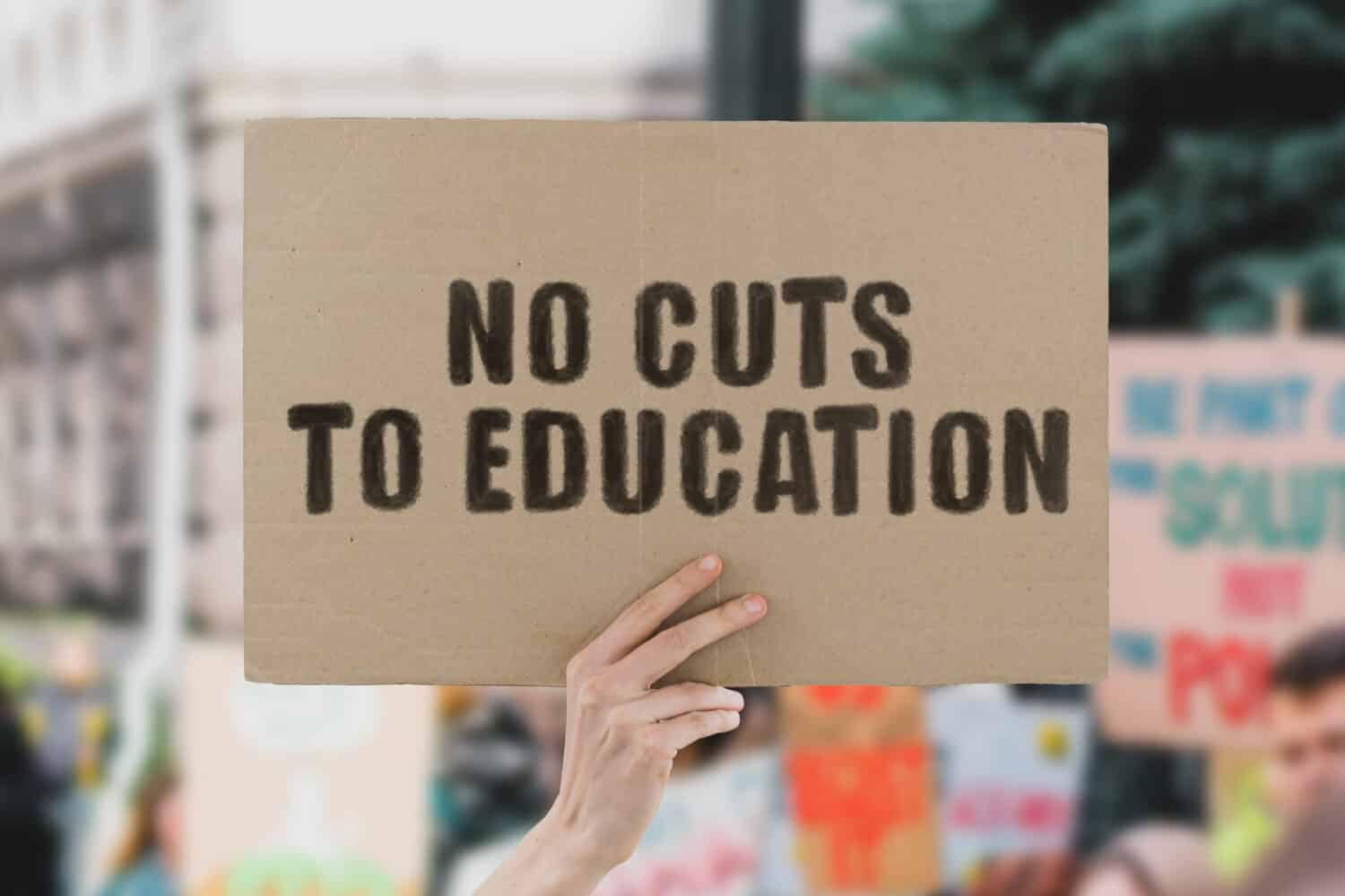 The phrase " No cuts to education " on a banner in men's hand. Human holds a cardboard with an inscription. Student. Children. Study. Changes. Teachers. Government. Classroom. High school. Classes