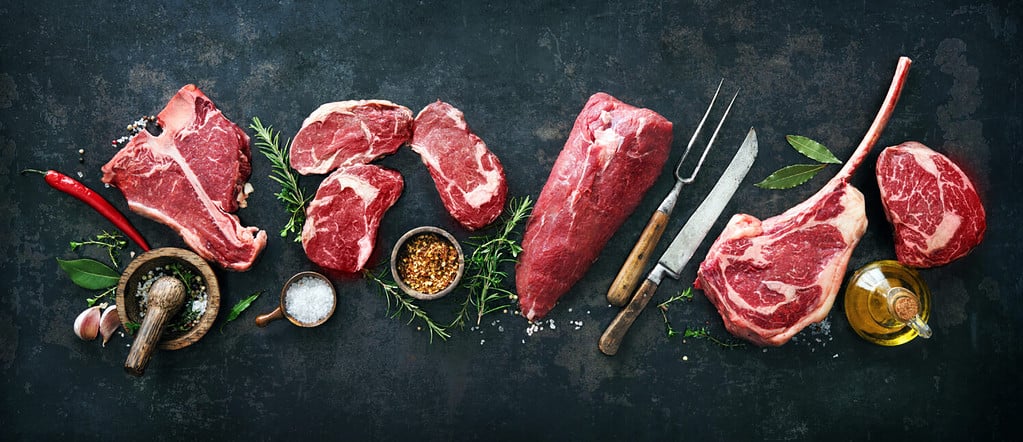 Variety of raw beef meat steaks for grilling with seasoning and utensils on dark rustic board