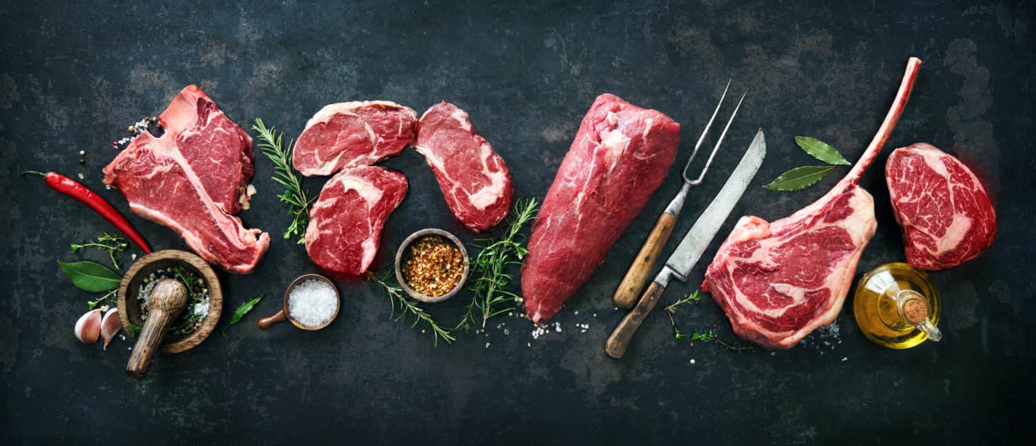 Variety of raw beef meat steaks for grilling with seasoning and utensils on dark rustic board