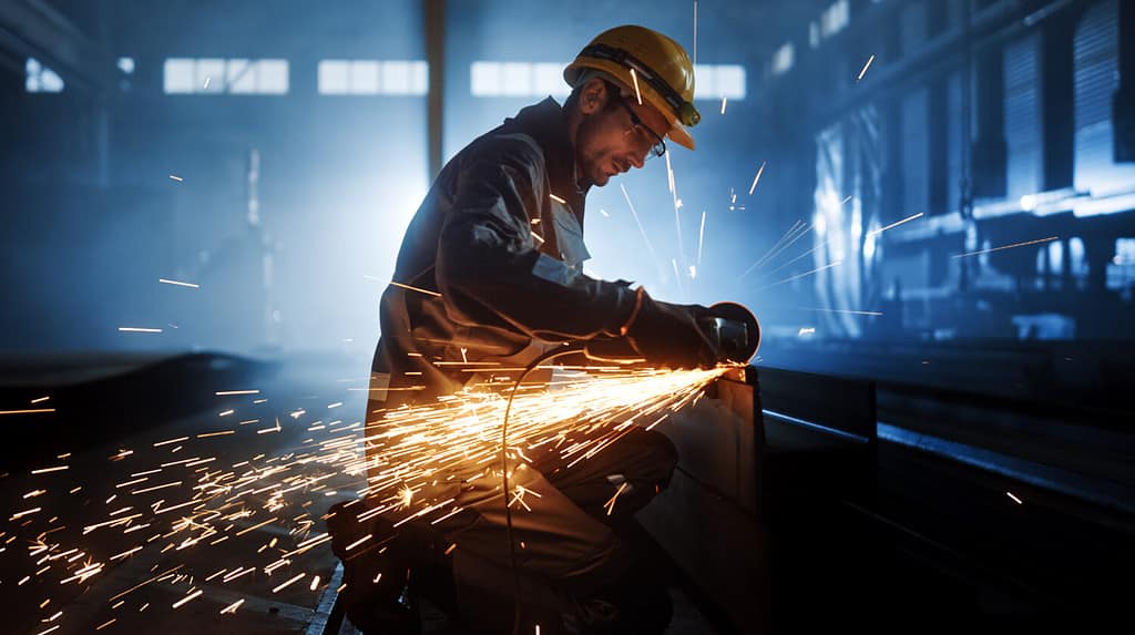 Heavy Industry Engineering Factory Interior with Industrial Worker Using Angle Grinder and Cutting a Metal Tube. Contractor in Safety Uniform and Hard Hat Manufacturing Metal Structures.