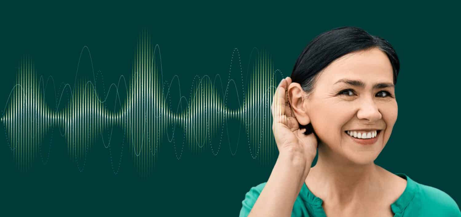 Positive woman with hand near ear and with luminous sound wave on green background showing variety of sounds going to her ear. Auditory health concept