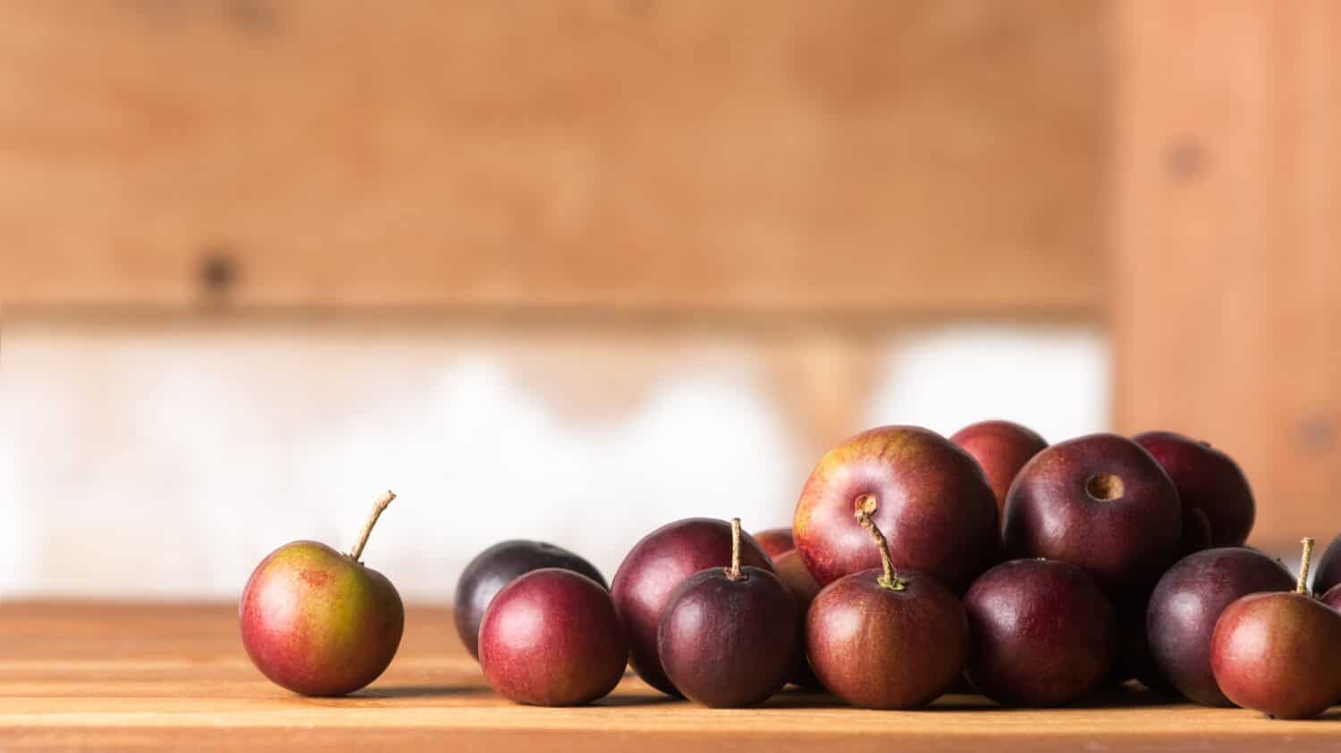 close-up of governor's plum fruits on wooden table top, flacourtia indica, also known as ramontchi, madagascar plum or indian plum, reddish black fleshy fruits, soft-focus wooden background