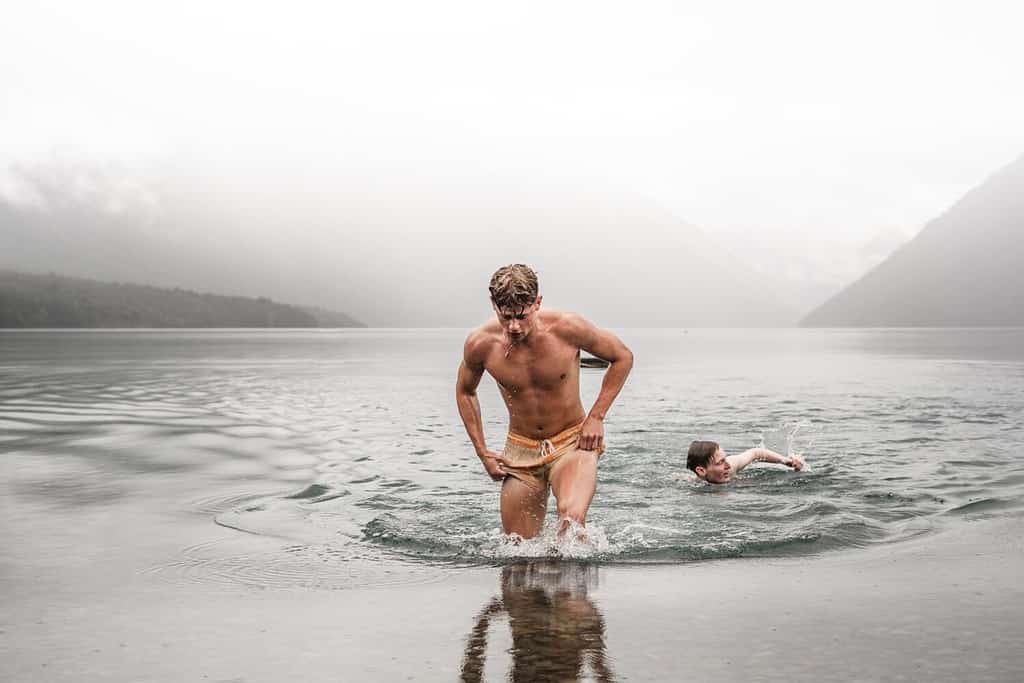 sporty caucasian boy without clothes and with a swimsuit getting out of the water wet and tired with his friend swimming in the cold waters of the lake on a cloudy day with a lot of fog, nelson lakes