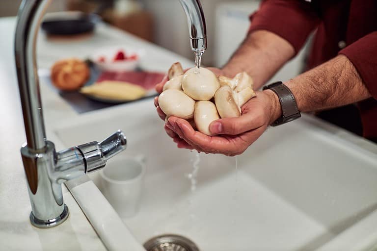The unknown man holding a fresh mushroom and cleaning them under a splash in the kitchen sink.