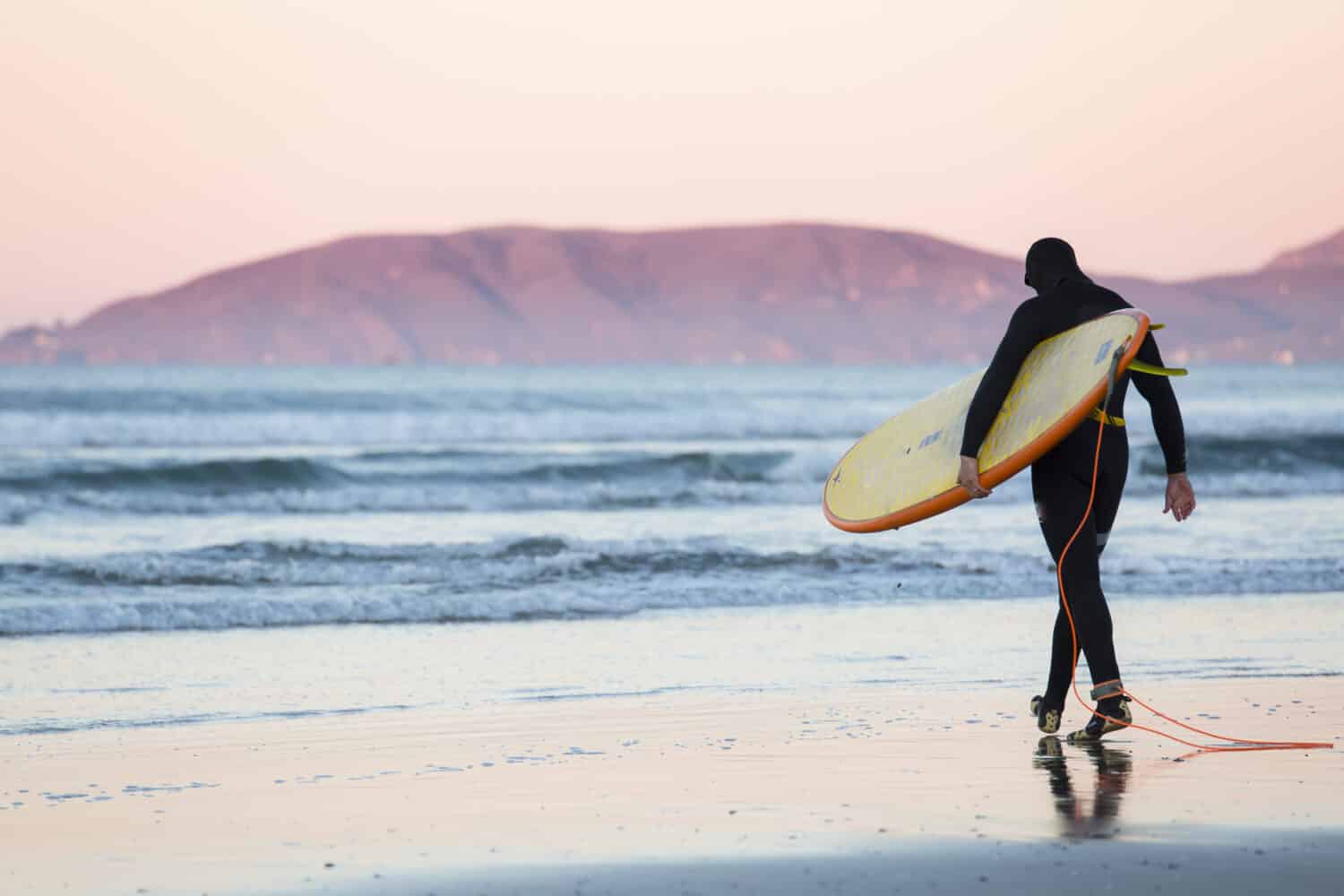 Surfer with surfboard and wetsuit walks towards the water at sunrise in Pismo Beach