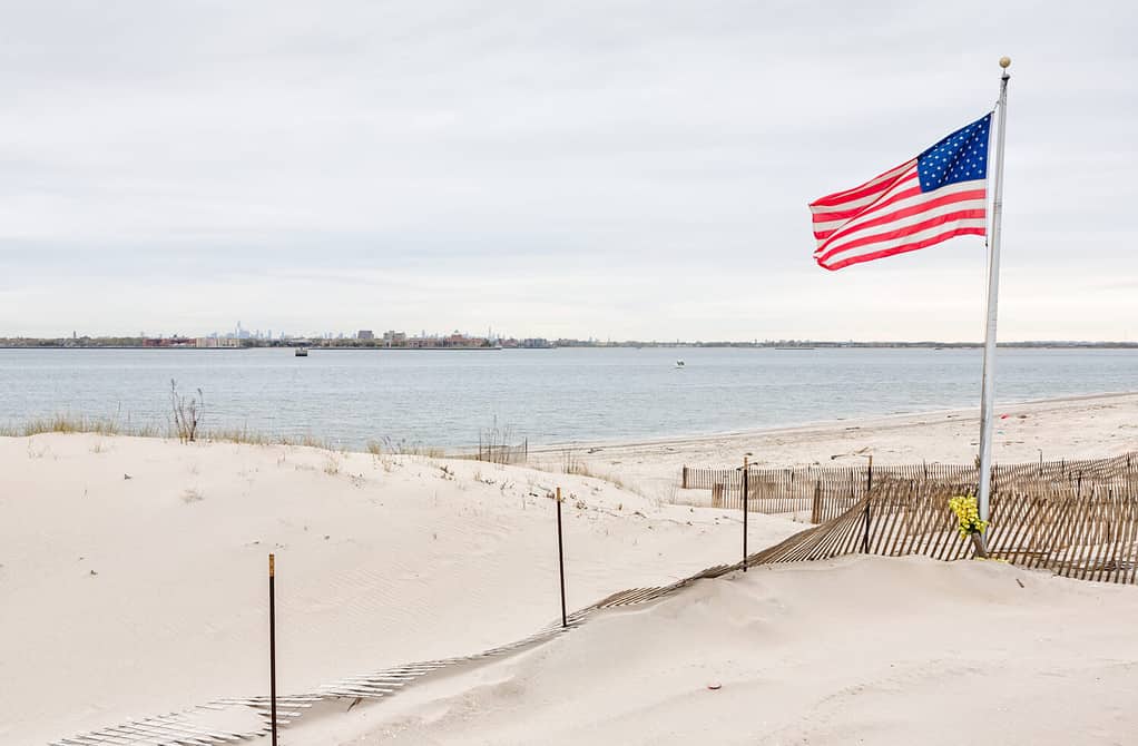 American flag on the beach of Breezy Point. Breezy Point is a neighborhood in the New York City borough of Queens, located on the western end of the Rockaway peninsula. Manhattan in the background
