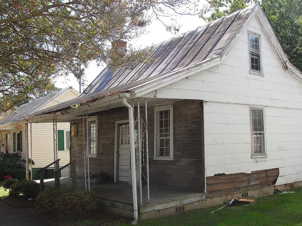 An old wooden house with a porch and white siding sits on a patch of grass.