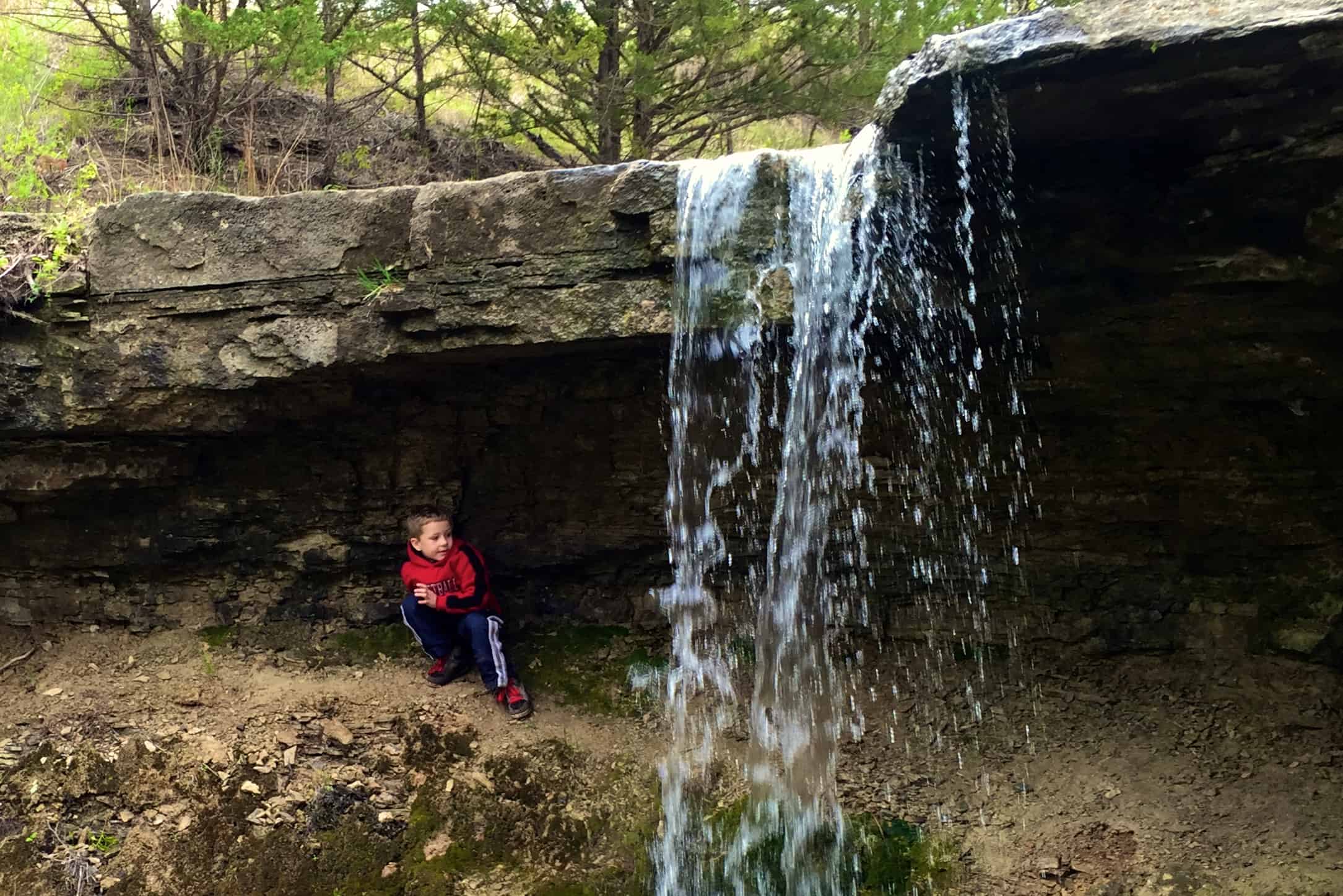 A small waterfall flows over a rock and a boy sits underneath it.
