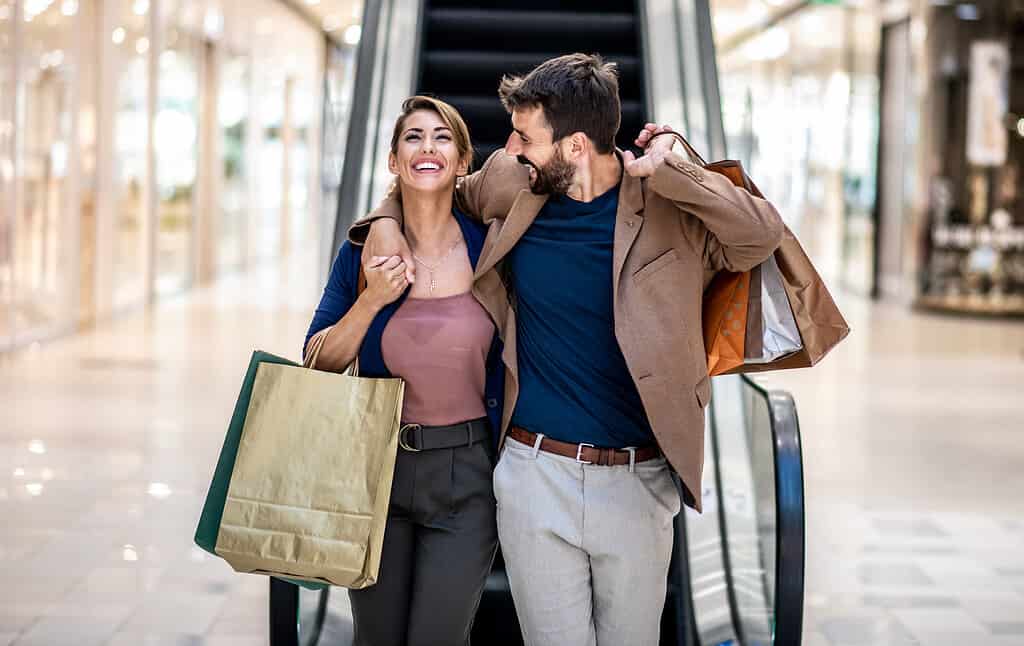 Young attractive happy couple hugging, smiling and holding shopping bags while walking in shopping mall.