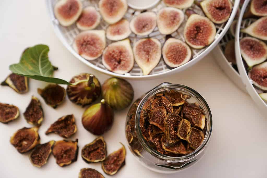 Fresh figs lie on the table next to dried figs in a glass jar and a dehydrator. Top view