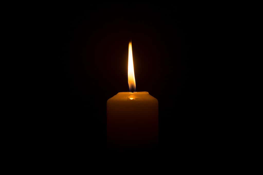 A single burning candle flame or light glowing on a white candle on black or dark background on table in church for Christmas, funeral or memorial service