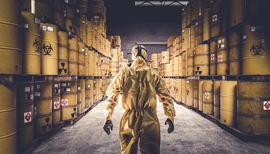 Man in yellow protective suit and gas mask, warehouse full of yellow metal barrels