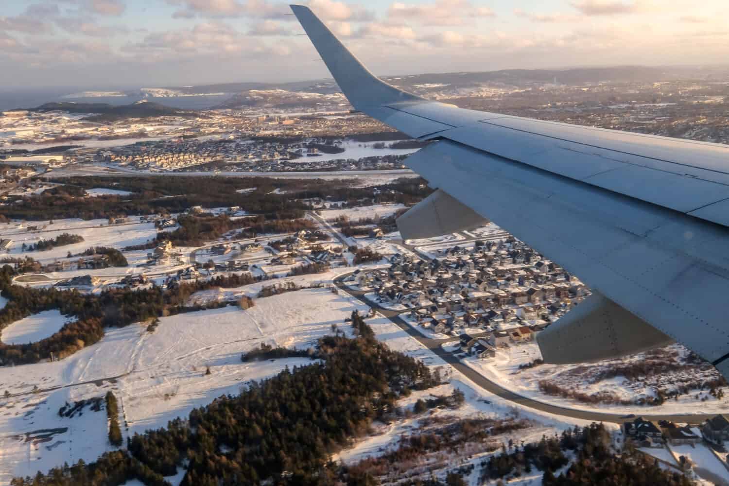 The left side wing of a large white commercial plane flying over the City of St. John's, Newfoundland under the evening sun. The sky is cloudy. The neighbourhood grounds below are covered in snow.