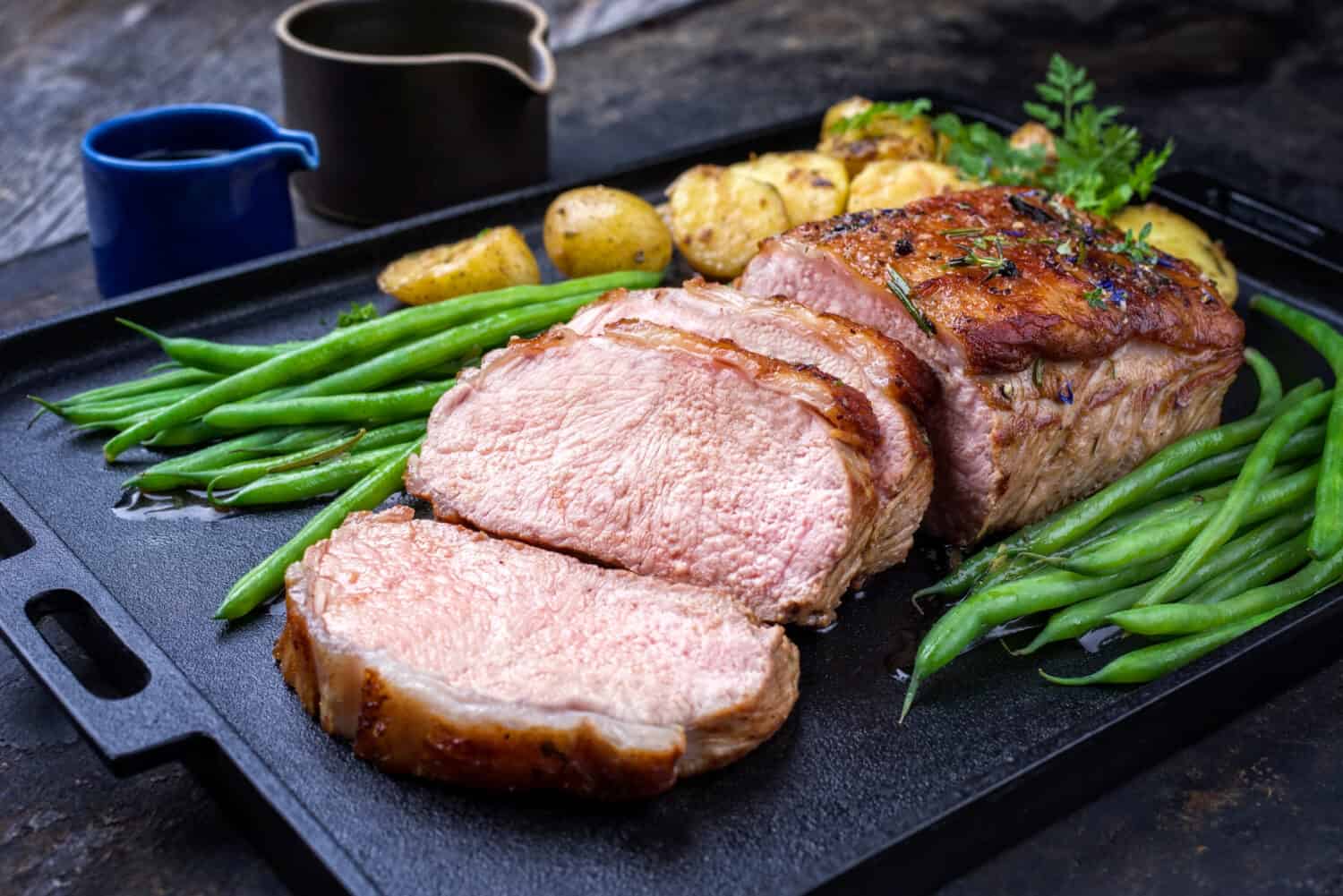 Traditional roasted dry aged veal tenderloin with beans and potatoes offered as closeup on a modern design cast iron tray 