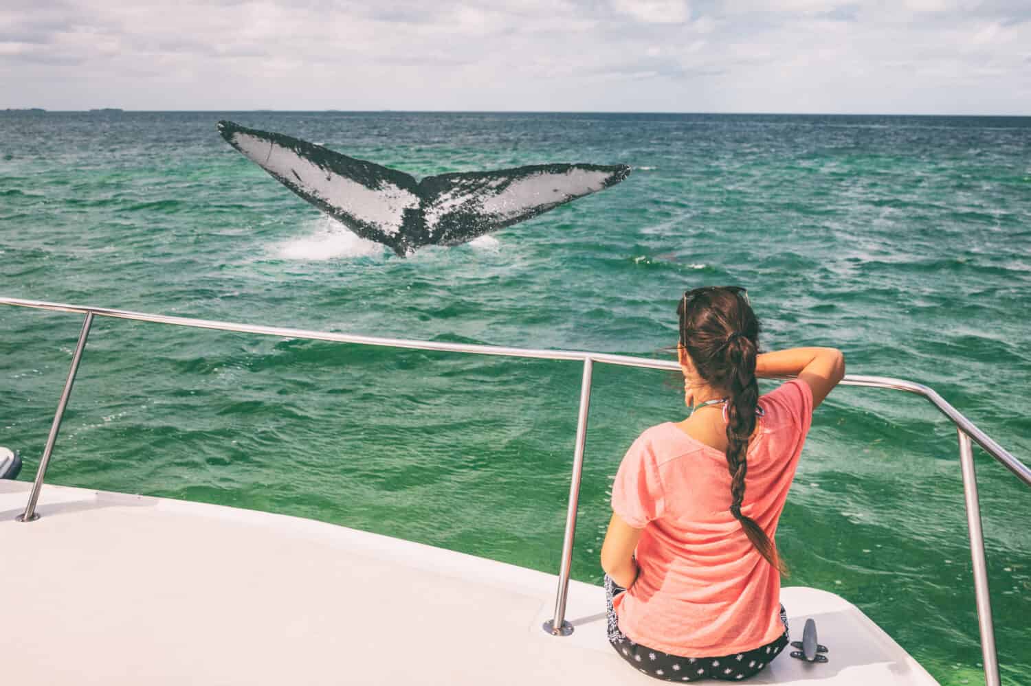 Whale watching boat tour tourist woman relaxing looking at humpback breaching flapping tail travel destination, summer vacation on deck of catamaran.