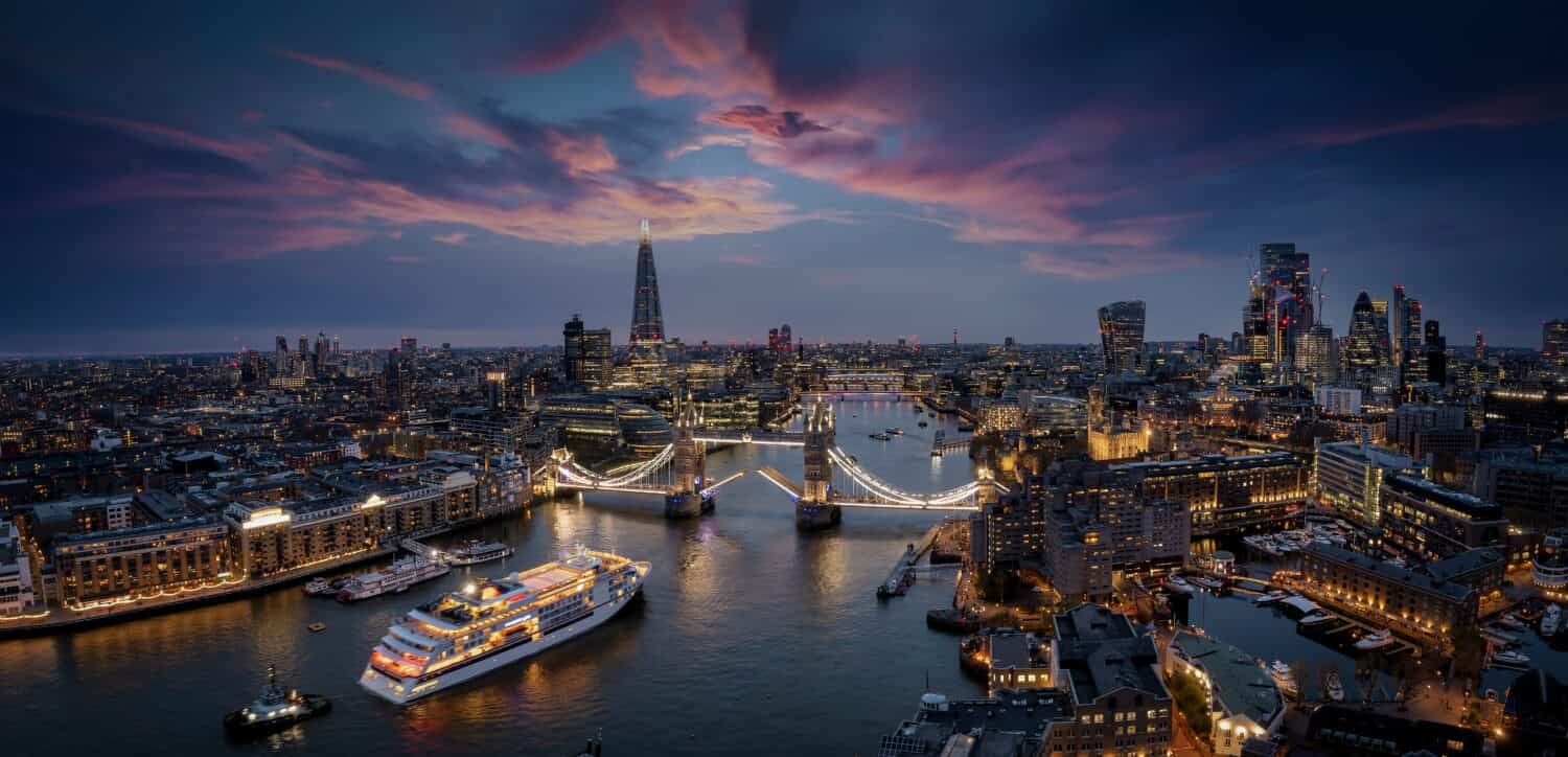 Panoramic, aerial view of the skyline of London with a motion blurred cruise ship passing under the lifted Tower Bridge during dusk, England
