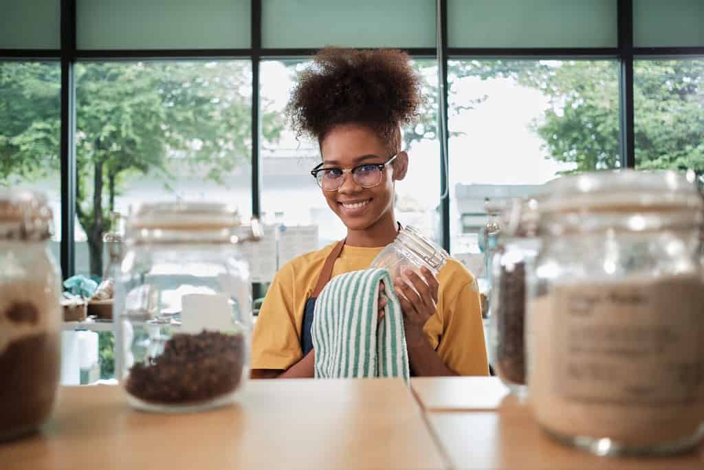 A young Black female shopkeeper cleans glass jars of natural organic products in reusable containers at a refill store, zero waste, a plastic-free grocery shop, and an eco-friendly retail business.