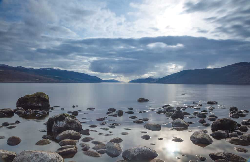 A view across Loch Ness looking down the length of the lake with rocks in the foreground and dark clouds above, in Scotland