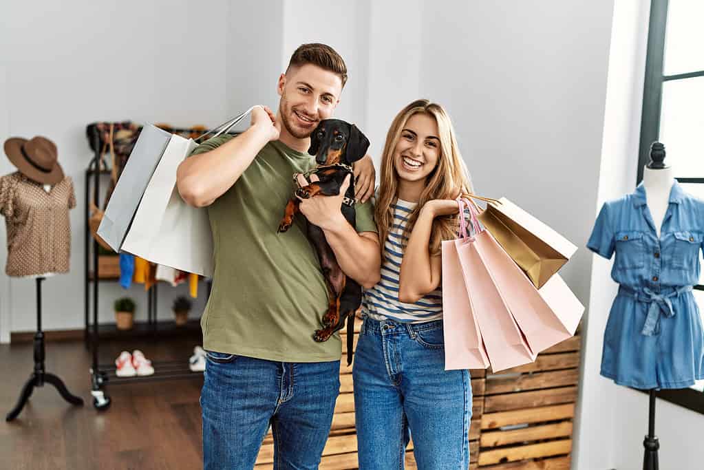 Young hispanic customer couple smiling happy holding shopping bags and dog at clothing store.