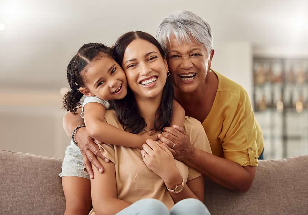 Grandmother, mom and child hug in a portrait for mothers day on a house sofa as a happy family in Colombia. Smile, mama and elderly woman love hugging young girl or kid and enjoying quality time