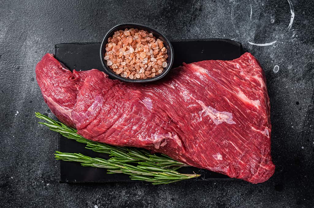 Raw American Tri Tip beef steak on marble board. Black background. Top view