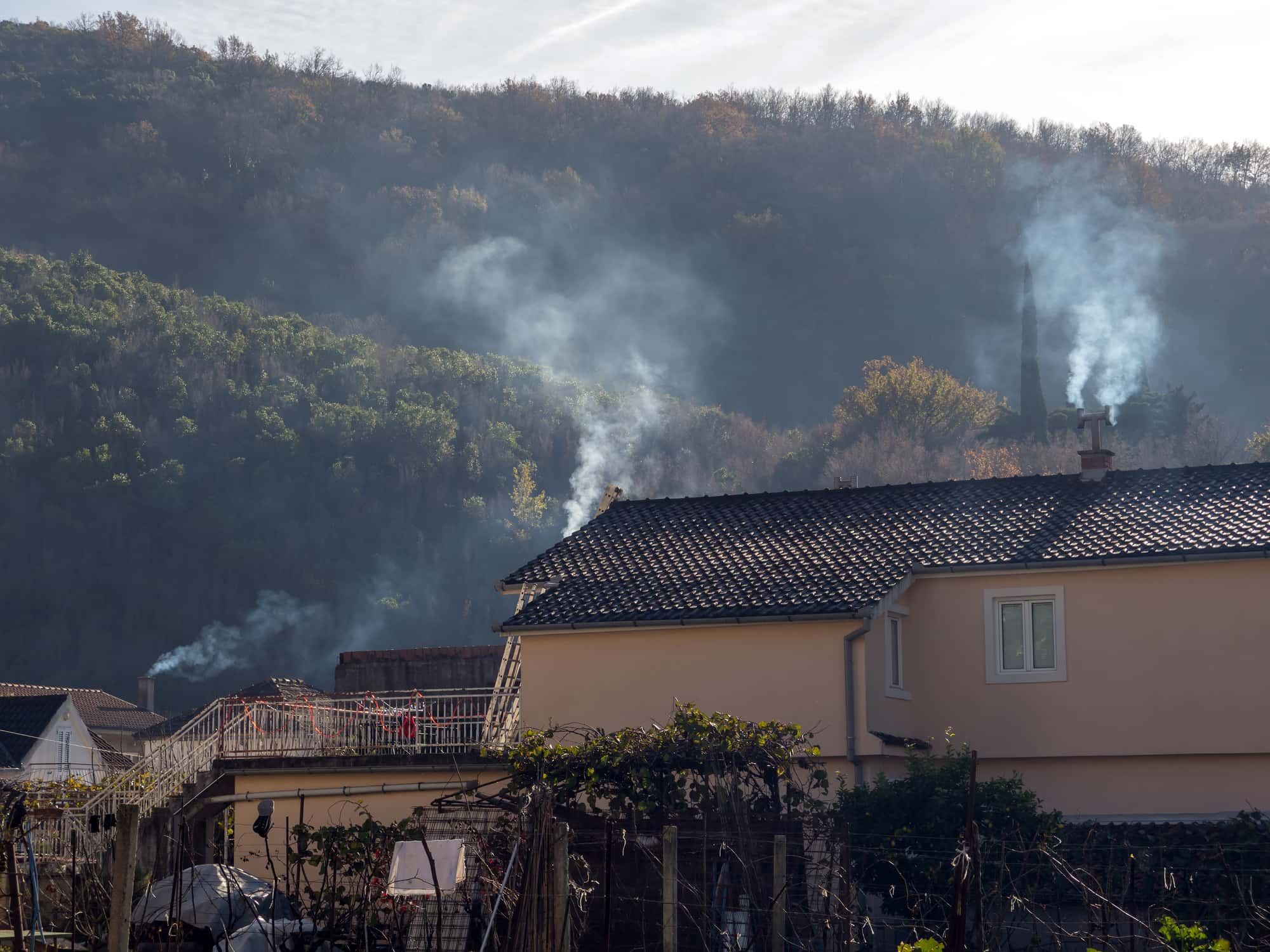 Smoke from houses chimneys of small town with tile roofs and hillside green trees at background side view