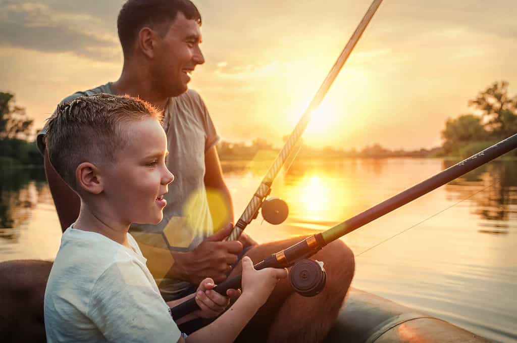Happy Father and Son together fishing from a boat at sunset time in summer day under beautiful sky on the lake.
