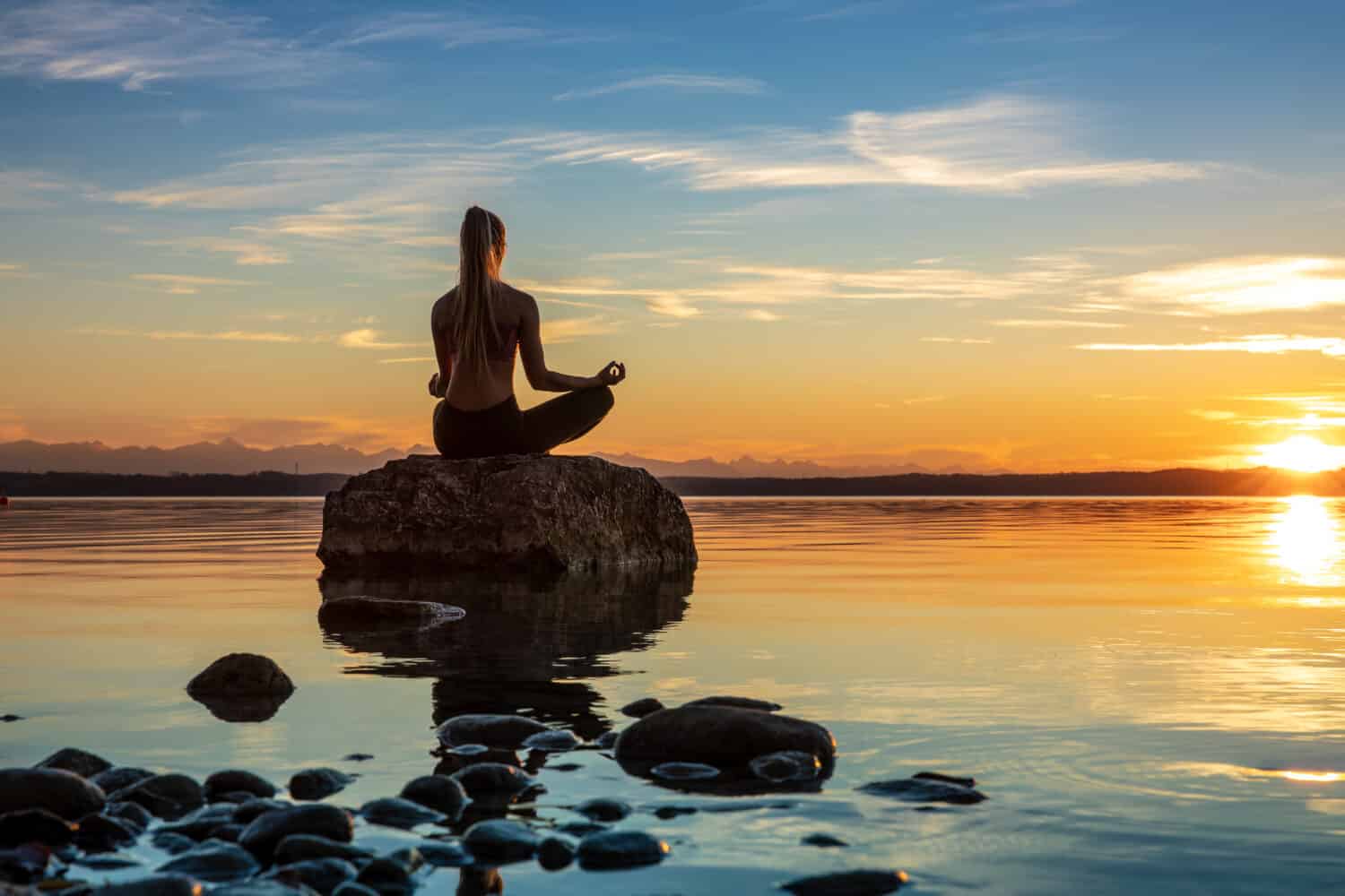 young beautiful woman girl are doing sport fitness yoga position on a stone at a beautiful lake, sea, see in the water - in the background you can see a colorful sundown - stone, water, orange sundown