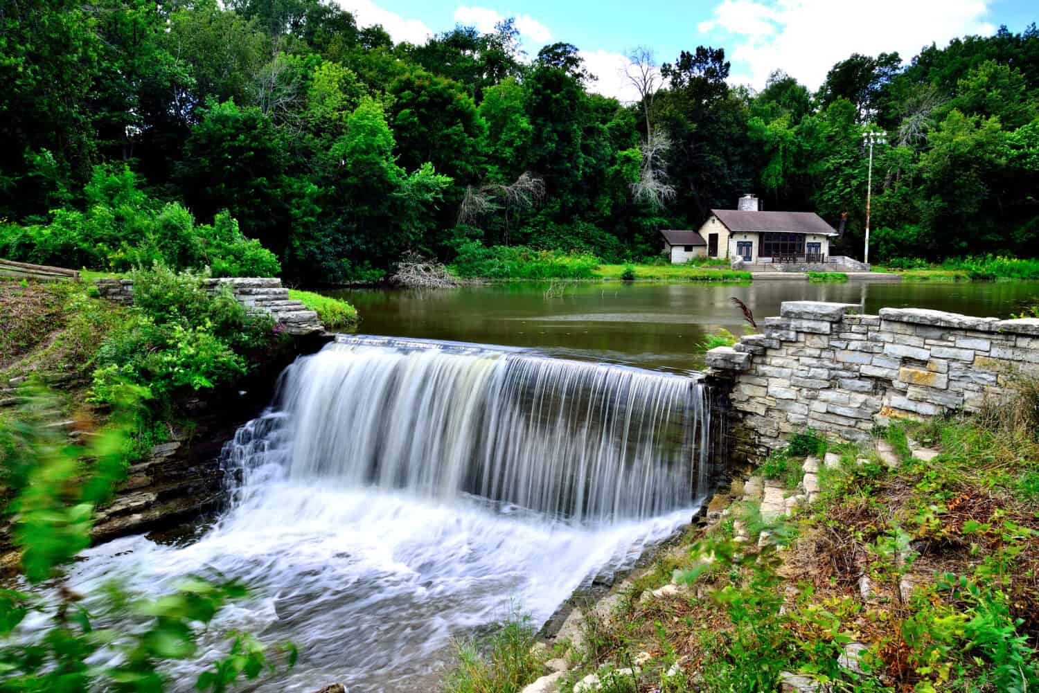 The dam at Oak Creek Pond with a park building in the background on a beautiful summer weekday.  The long exposure gives the waterfall a cottonlike appearance.  The park is in South Milwaukee.  