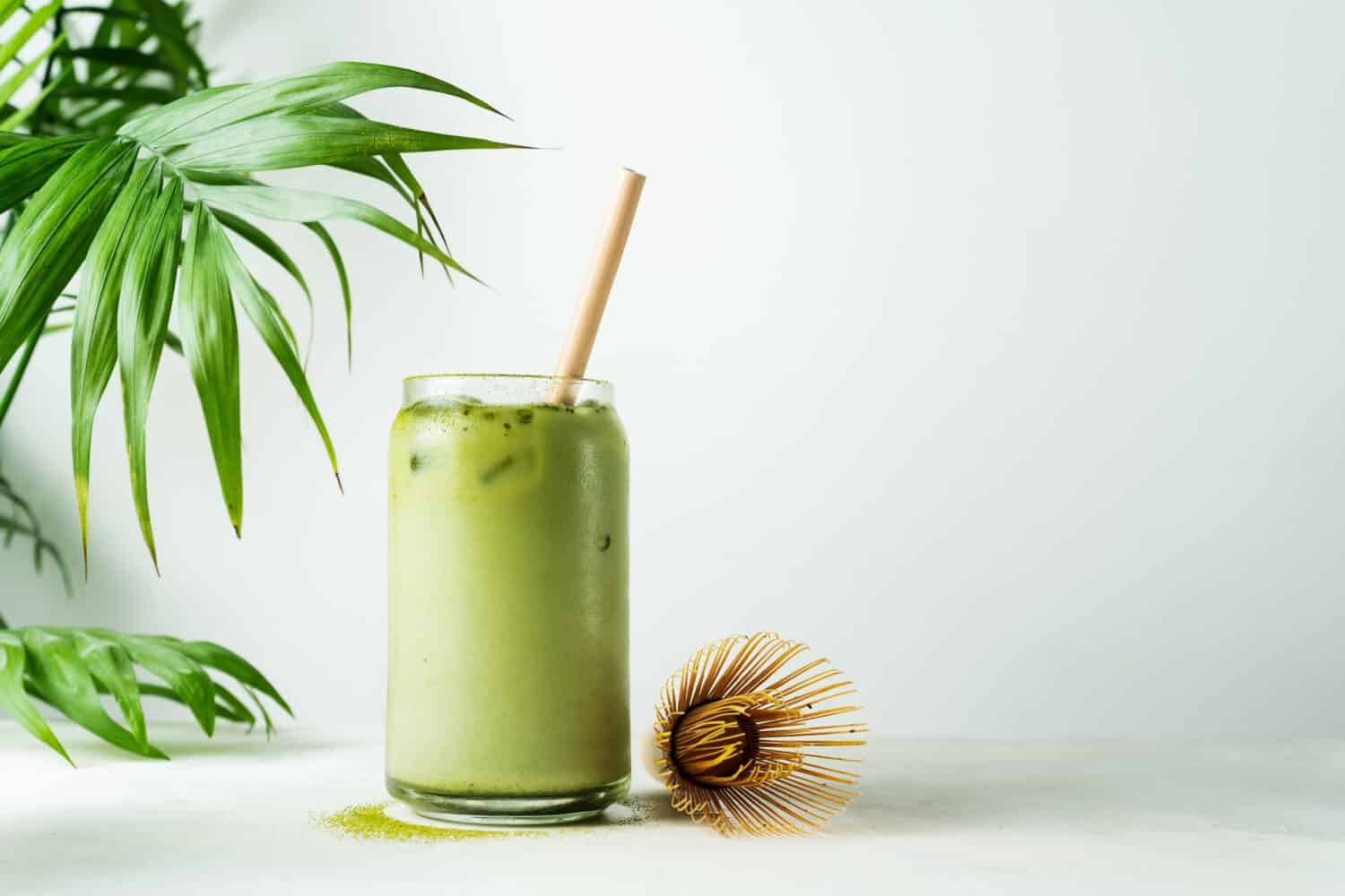 Making Japanese iced matcha latte, green tea with milk, soy milk, traditional matcha tools, with bamboo straw in glass on white background.