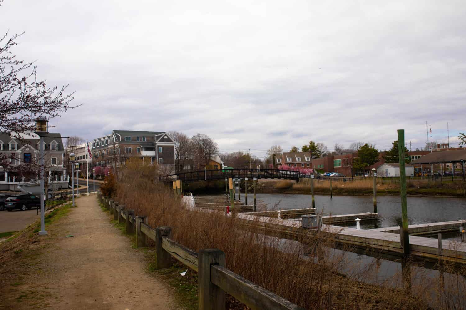Small town landscape - Milford marina on the right, walking path on the left,  colorful townhouses, and walking bridge in the back on a grey gloomy day in Milford, CT. 
