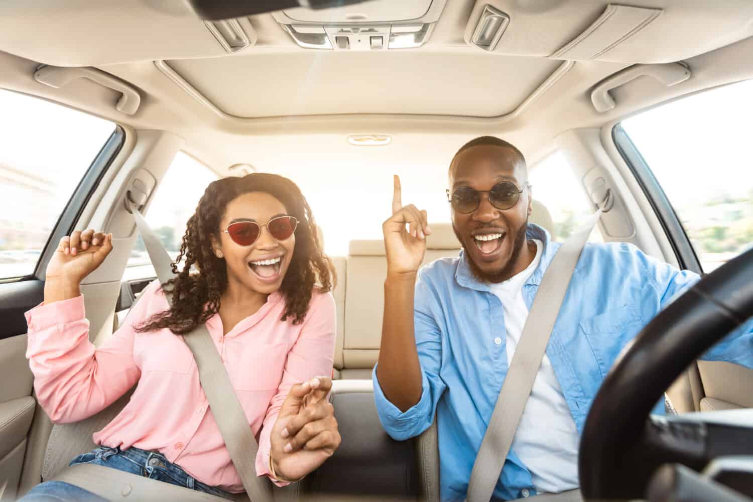 We Are Going On Vacation. Frontal view portrait of cheerful positive African American guy and lady in sunglasses driving their automobile, dancing to music and singing favorite song, looking at camera