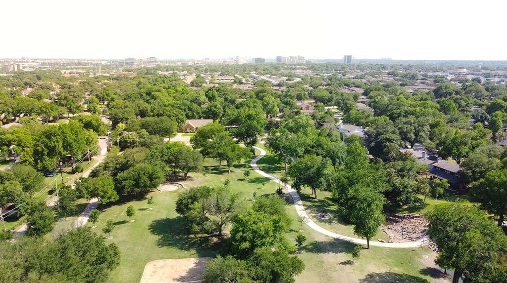 Lush green grassy park with trail system and matured trees near established residential neighborhood with downtown Richardson in background. Aerial view subdivision sprawl in North Texas, USA
