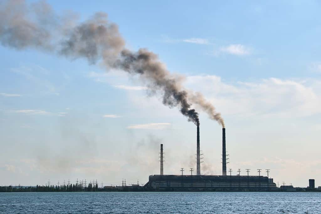 Coal power plant high pipes with black smoke moving upwards polluting atmosphere over lake water
